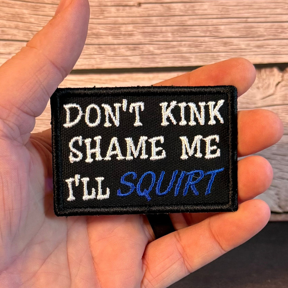 A hand holding a rectangular black embroidered patch with white and blue text that reads 'DON'T KINK SHAME ME I'LL SQUIRT' against a rustic wood plank background.