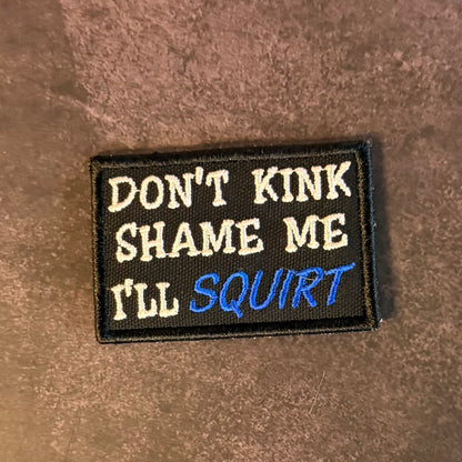 A rectangular embroidered patch with the text 'DON'T KINK SHAME ME I'LL SQUIRT' in white and blue lettering on a black background, placed on a brown textured surface.