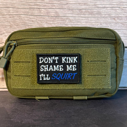 An olive green tactical pouch with a black rectangular patch featuring the text 'DON'T KINK SHAME ME I'LL SQUIRT' in white and blue embroidery, set against a light wood background.