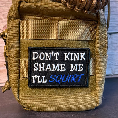 A close-up of a tan tactical pouch featuring a black embroidered patch with the text 'DON'T KINK SHAME ME I'LL SQUIRT' in white and blue lettering.