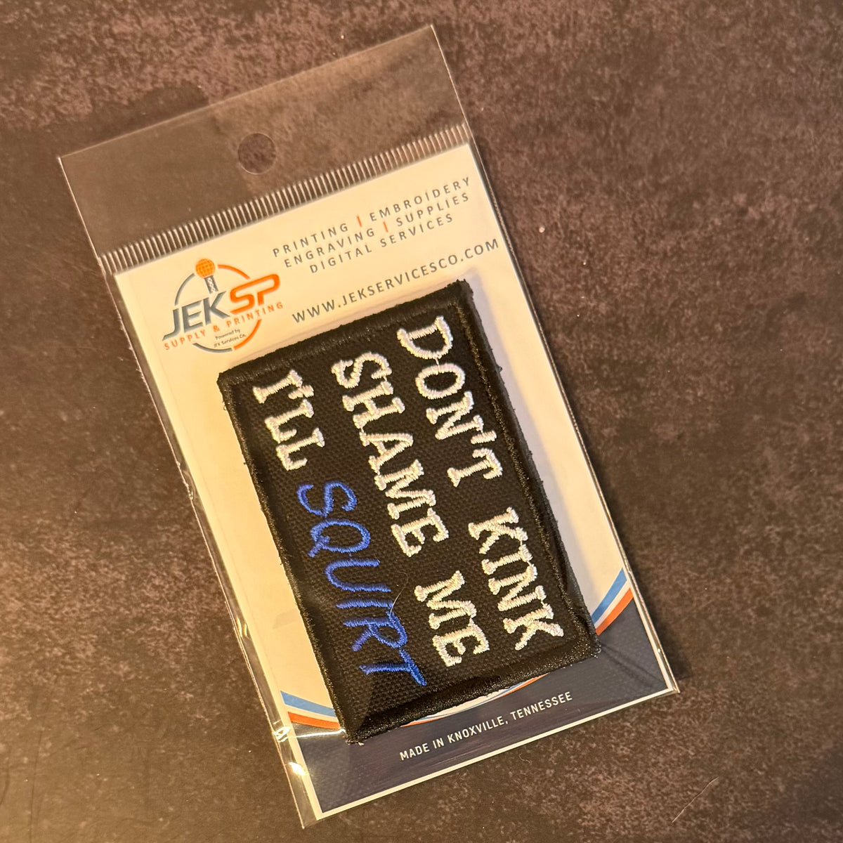 A black embroidered patch with the text 'DON'T KINK I'LL SCURT' in white and blue, sealed in a clear plastic sleeve with a branded card backing. The card shows the JEKSP brand and reads 'PRINTING | EMBROIDERY | ENGRAVING | SUPPLIES DIGITAL SERVICES'.