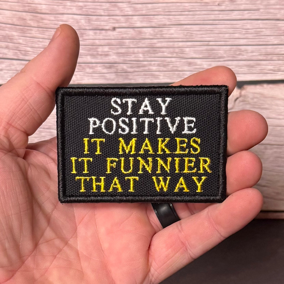 A hand holding a rectangular black embroidered patch with the text 'STAY POSITIVE IT MAKES IT FUNNIER THAT WAY' in white and yellow lettering, against a light wood plank background.