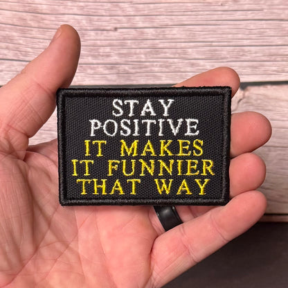 A hand holding a rectangular black embroidered patch with the text 'STAY POSITIVE IT MAKES IT FUNNIER THAT WAY' in white and yellow lettering, against a light wood plank background.