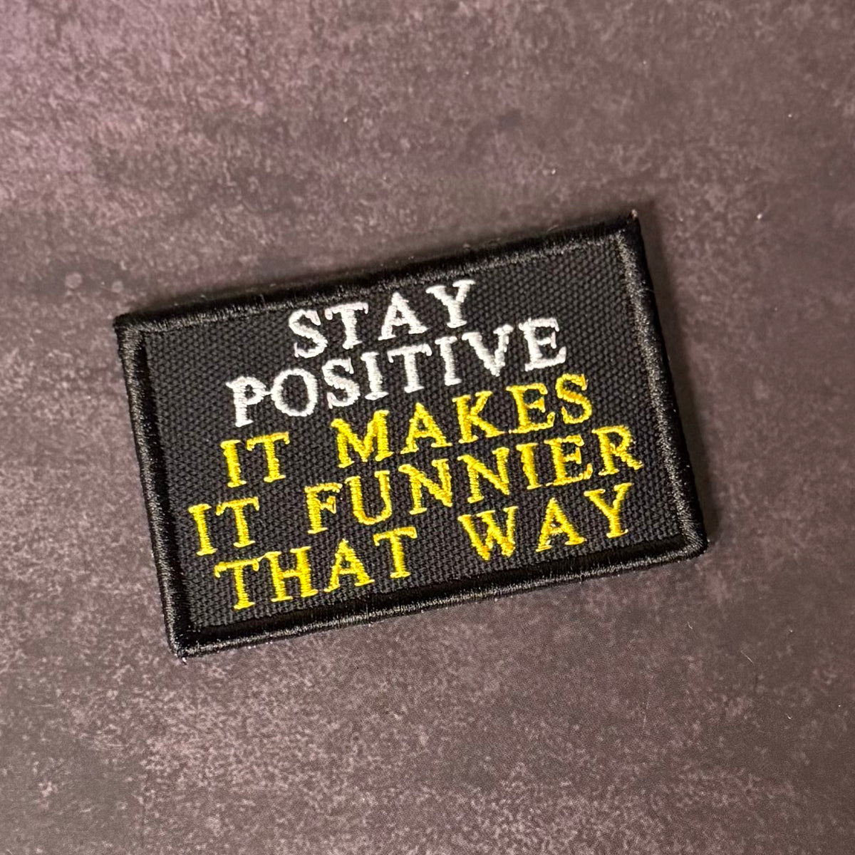 A rectangular black embroidered patch with the text 'STAY POSITIVE IT MAKES IT FUNNIER THAT WAY' in white and yellow lettering, placed on a textured dark brown surface.