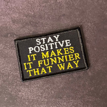 A rectangular black embroidered patch with the text 'STAY POSITIVE IT MAKES IT FUNNIER THAT WAY' in white and yellow lettering, placed on a textured dark brown surface.