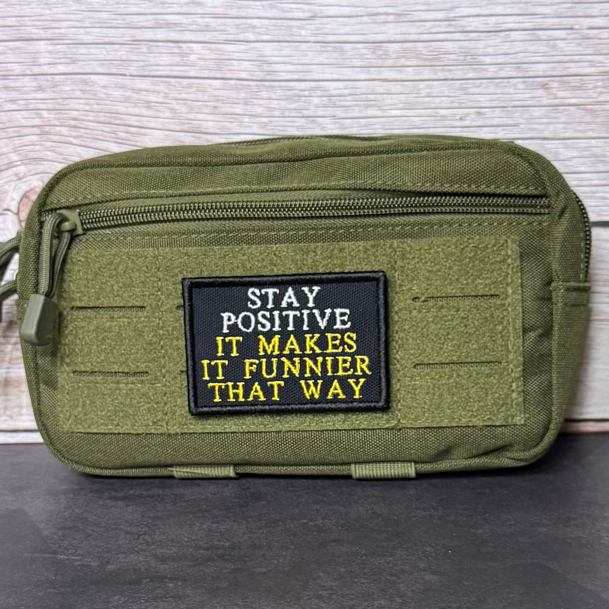 An olive green tactical pouch with a black rectangular patch featuring the text 'STAY POSITIVE IT MAKES IT FUNNIER THAT WAY' in white and yellow embroidery, set against a light wood background.