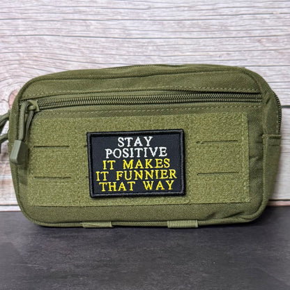 An olive green tactical pouch with a black rectangular patch featuring the text 'STAY POSITIVE IT MAKES IT FUNNIER THAT WAY' in white and yellow embroidery, set against a light wood background.