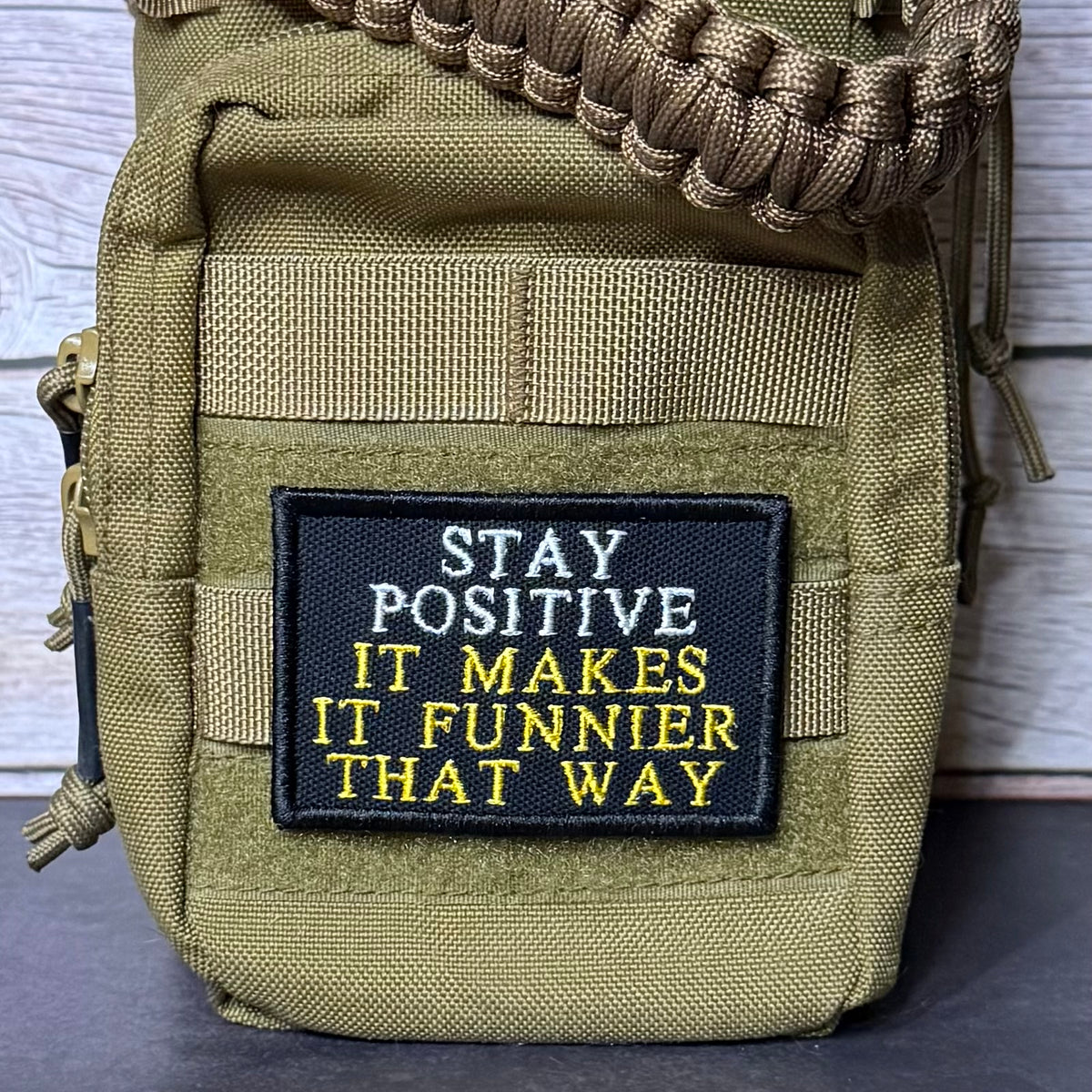 An olive green tactical pouch with a black embroidered patch that reads 'STAY POSITIVE IT MAKES IT FUNNIER THAT WAY' in white and yellow text. A brown paracord strap is threaded through the top loop, and the pouch is photographed against a light wood background.