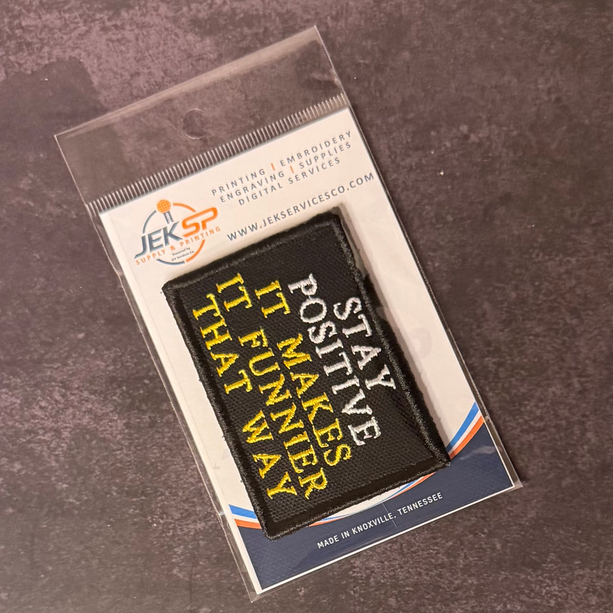 A black rectangular embroidered patch with yellow and white text reading 'STAY POSITIVE IT MAKES IT FUNNY THAT WAY' in a clear plastic sleeve on a branded card, photographed on a dark textured surface.