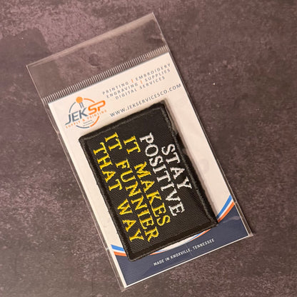 A black rectangular embroidered patch with yellow and white text reading 'STAY POSITIVE IT MAKES IT FUNNY THAT WAY' in a clear plastic sleeve on a branded card, photographed on a dark textured surface.
