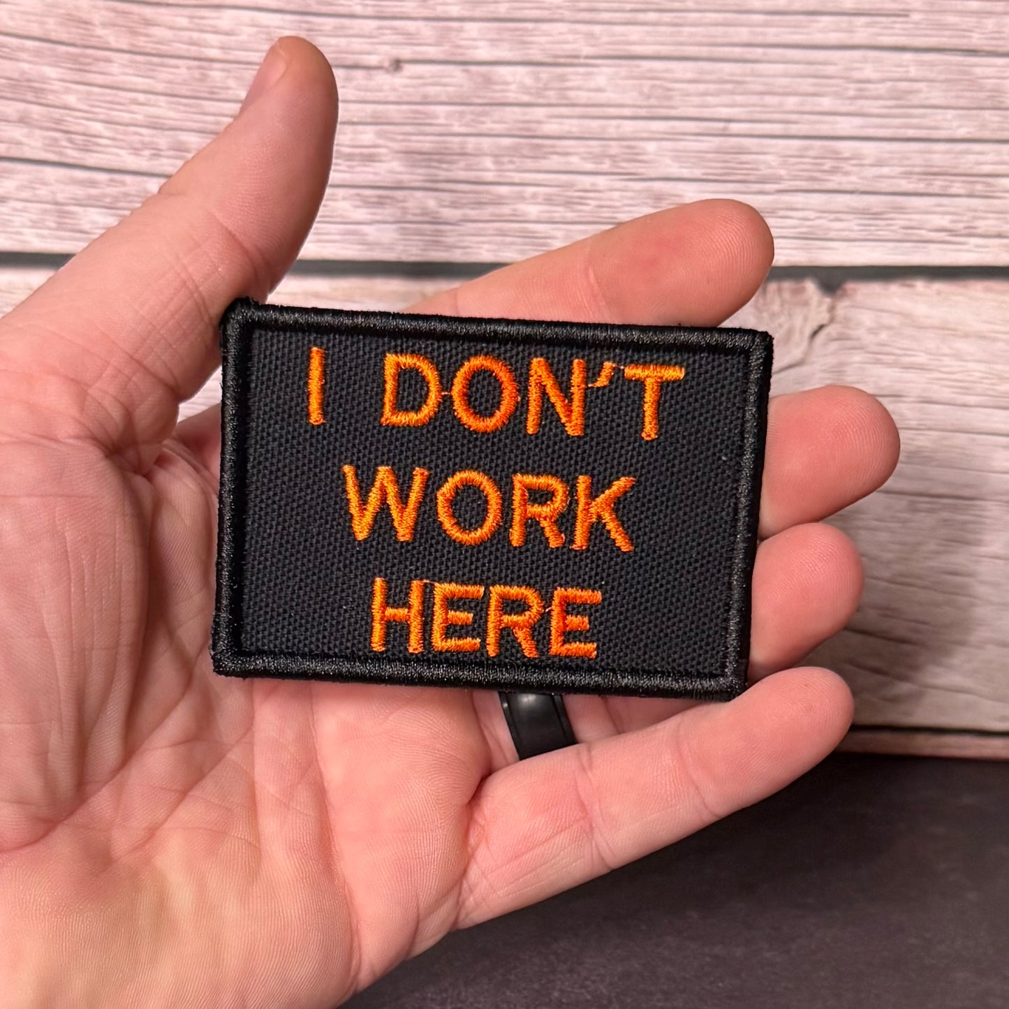 A hand holding a rectangular black embroidered patch with orange text that reads 'I DON'T WORK HERE' against a light wood plank background.