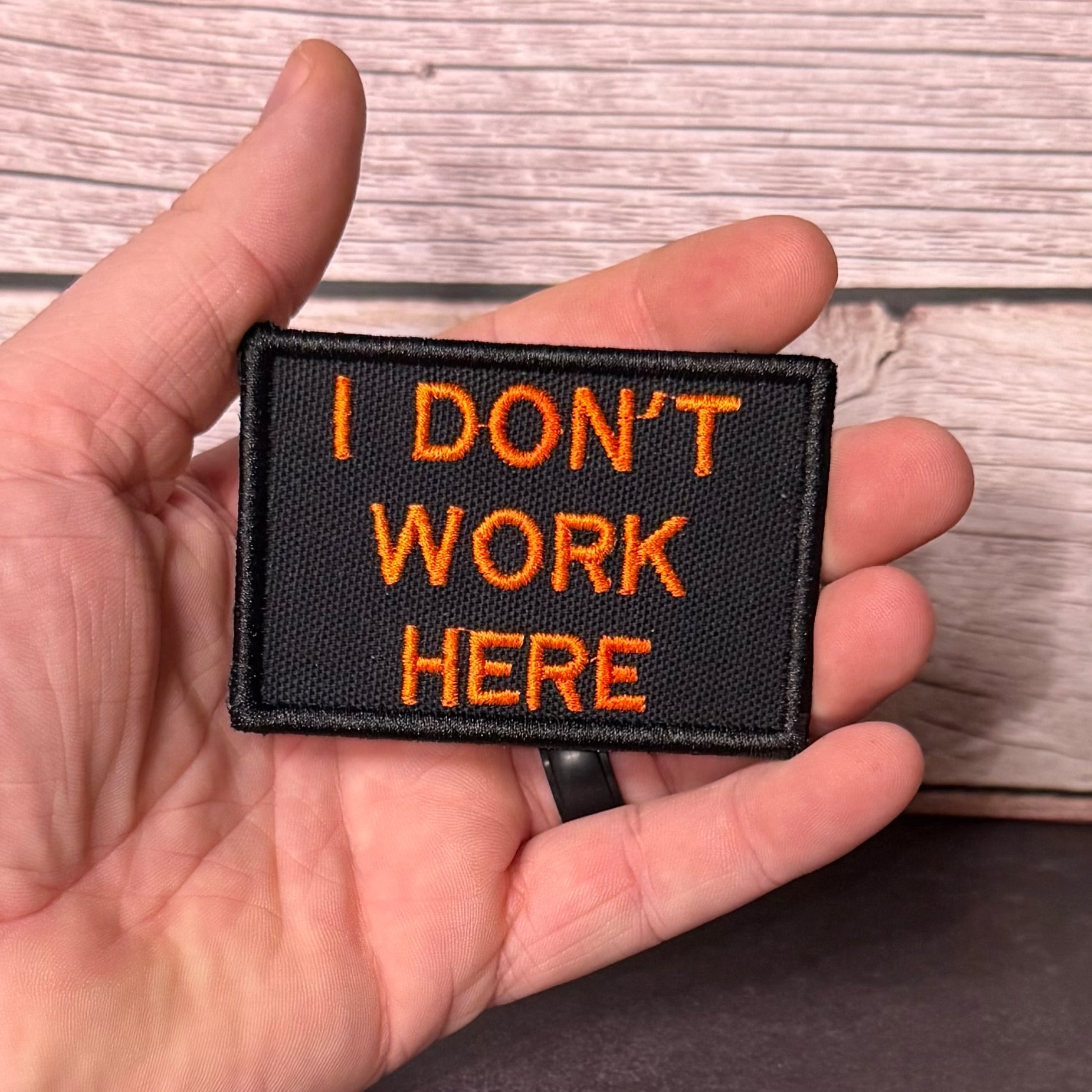 A hand holding a rectangular black embroidered patch with orange text that reads 'I DON'T WORK HERE' against a light wood plank background.