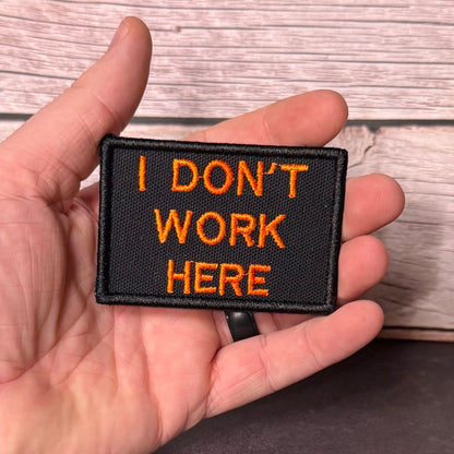A hand holding a rectangular black embroidered patch with orange text that reads 'I DON'T WORK HERE' against a light wood plank background.