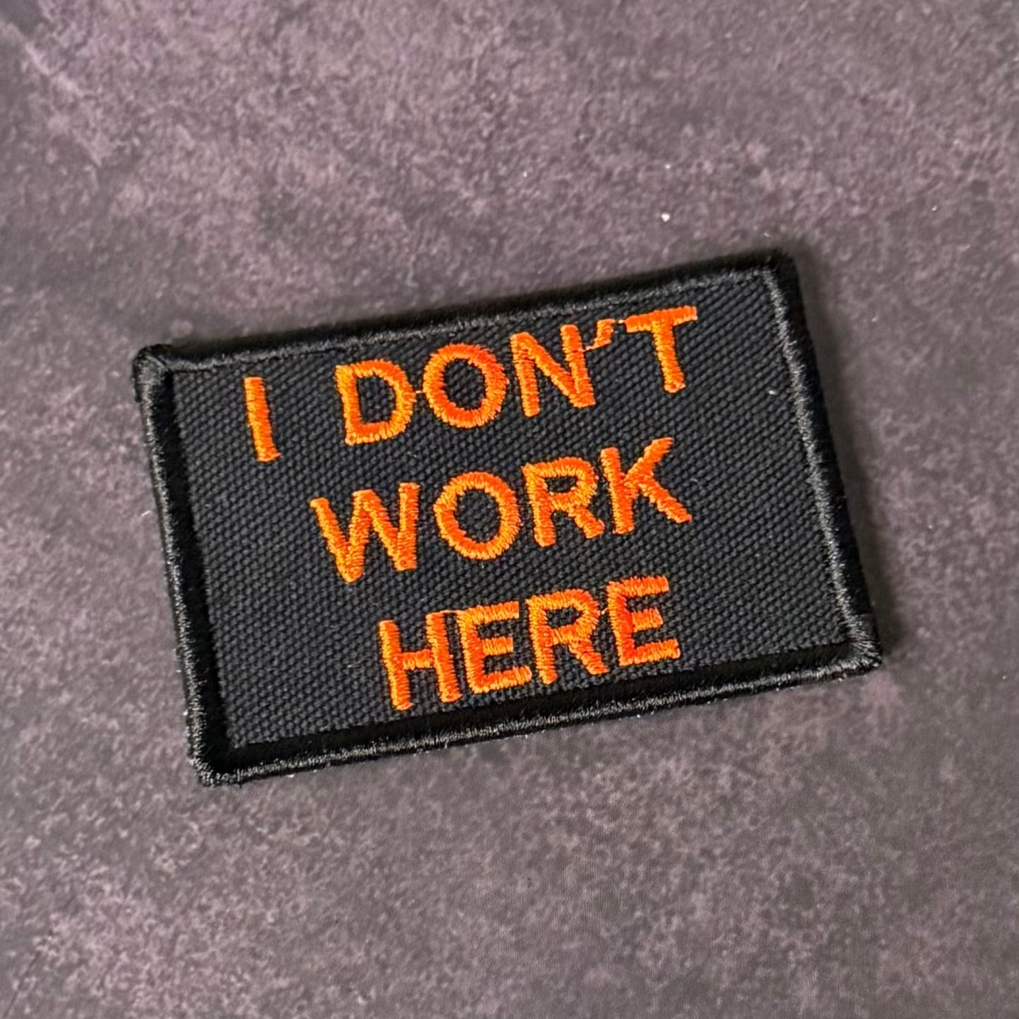 A rectangular black embroidered patch with orange text reading 'I DON'T WORK HERE' on a textured gray background.