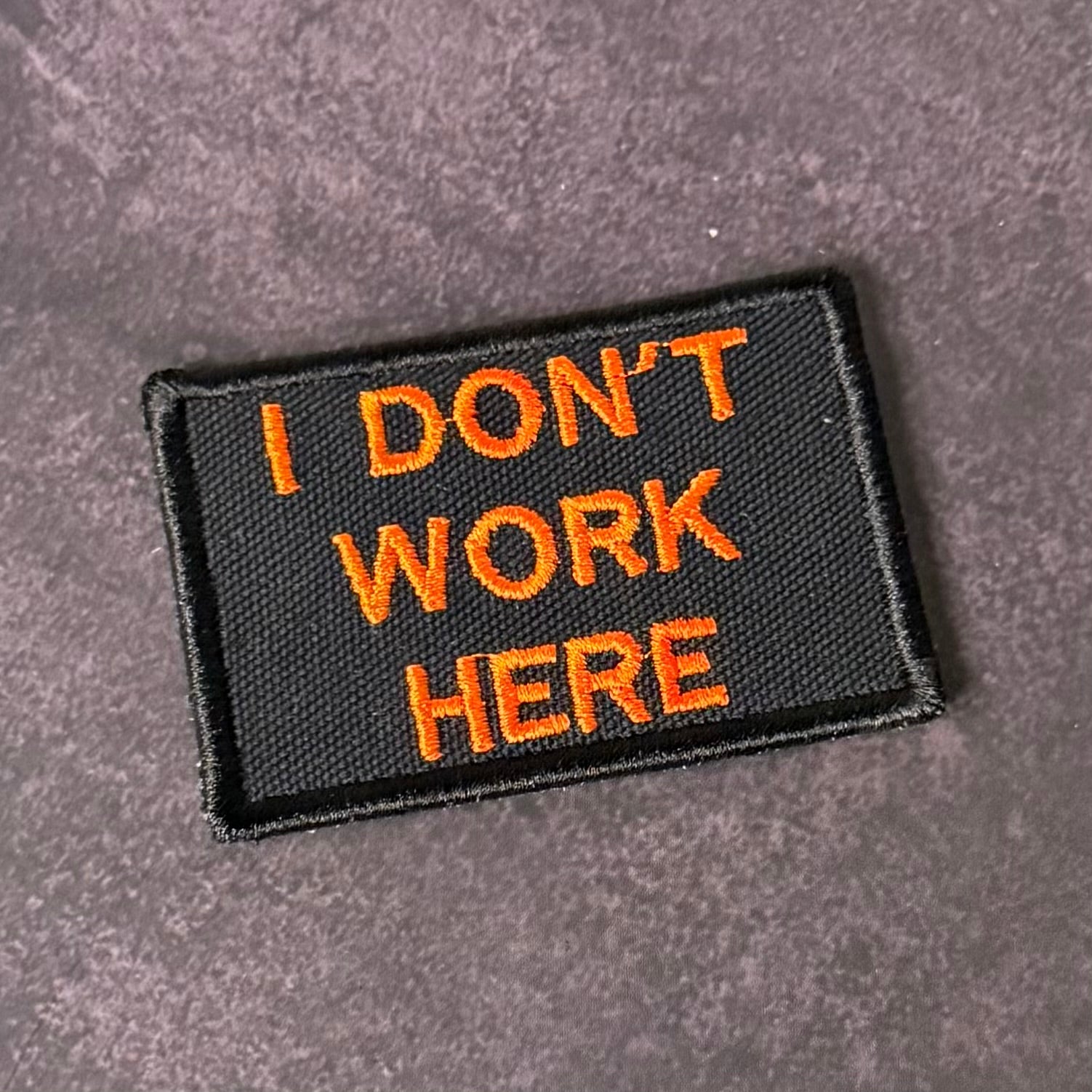 A rectangular black embroidered patch with orange text reading 'I DON'T WORK HERE' on a textured gray background.