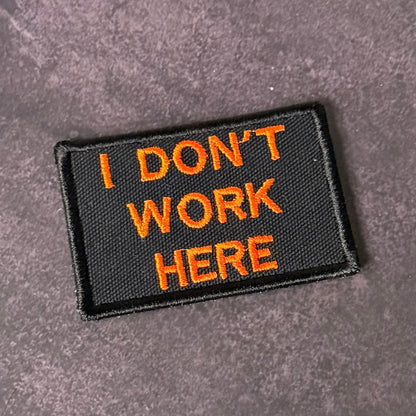 A rectangular black embroidered patch with orange text reading 'I DON'T WORK HERE' on a textured gray background.