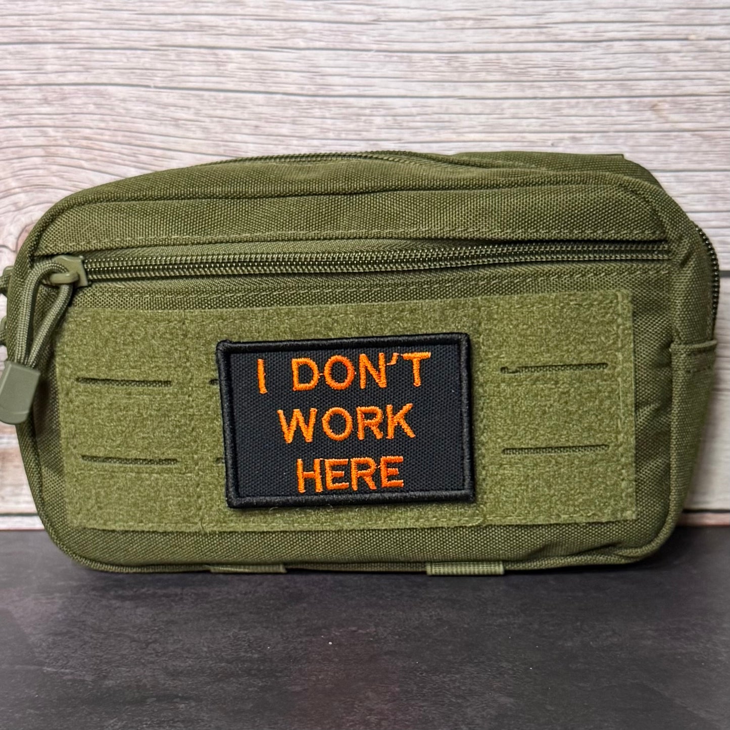 An olive green tactical pouch with a black rectangular patch featuring orange embroidered text that reads 'I DON'T WORK HERE'. The pouch is displayed on a dark surface against a light wood background.