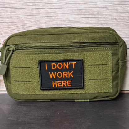 An olive green tactical pouch with a black rectangular patch featuring orange embroidered text that reads 'I DON'T WORK HERE'. The pouch is displayed on a dark surface against a light wood background.