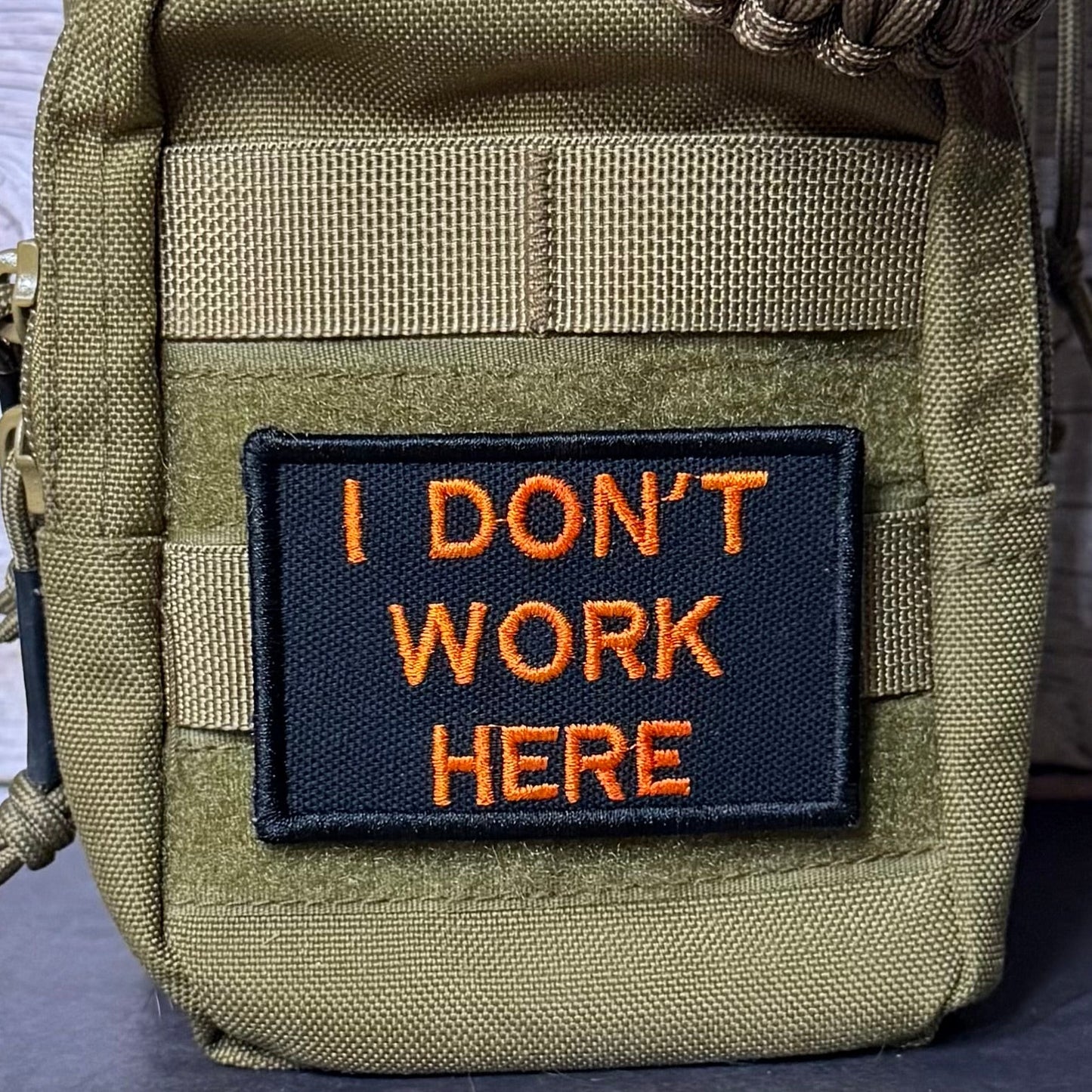 An olive green tactical pouch with a black rectangular patch featuring orange embroidered text that reads 'I DON'T WORK HERE'. The pouch has a woven strap and a paracord detail at the top, set against a rustic wood background.