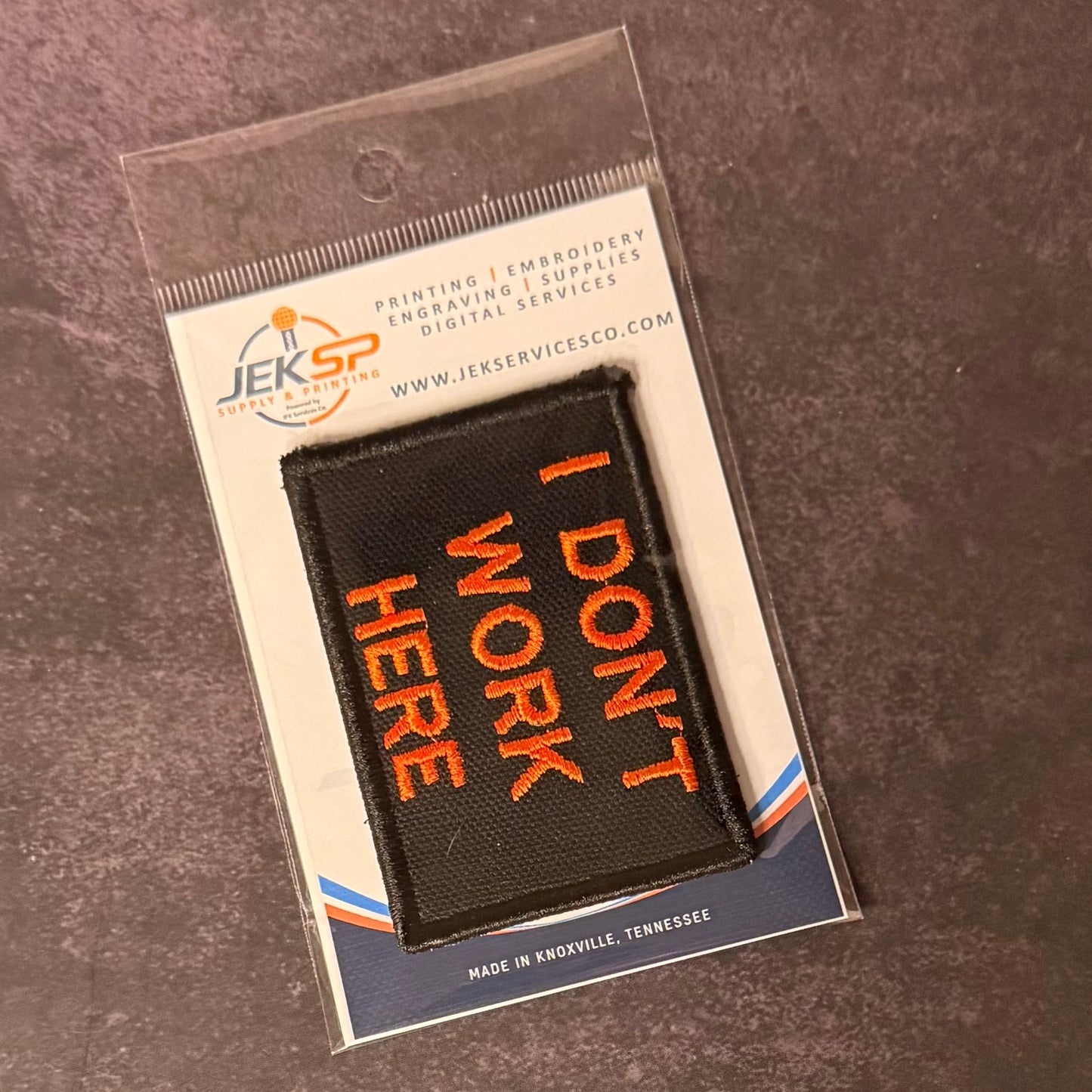 A black rectangular embroidered patch with orange text reading 'I DON'T WORK HERE' in a clear plastic sleeve, placed on a dark textured surface. The card backing shows the JEKSP brand and service details.