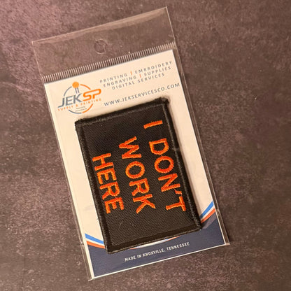 A black rectangular embroidered patch with orange text reading 'I DON'T WORK HERE' in a clear plastic sleeve, placed on a dark textured surface. The card backing shows the JEKSP brand and service details.