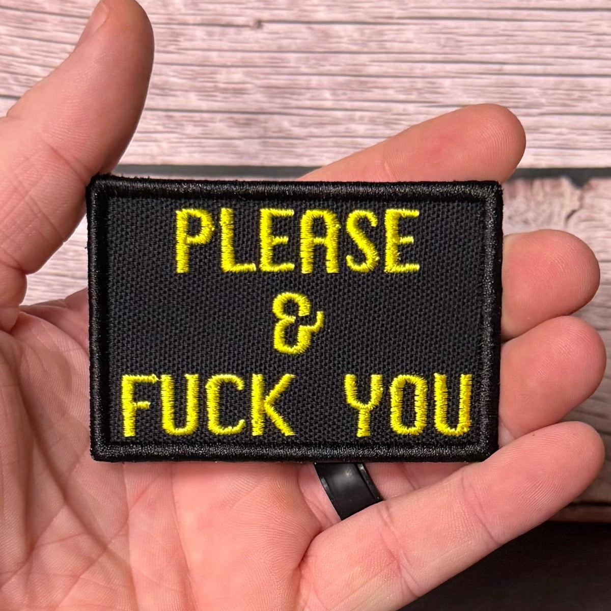 A hand holding a rectangular black embroidered patch with yellow text that reads 'PLEASE & FUCK YOU' against a light wood plank background.