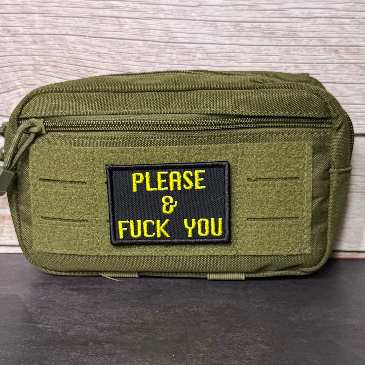 An olive green tactical pouch with a black rectangular patch featuring yellow embroidered text that reads 'PLEASE & FUCK YOU', set against a light wood background.