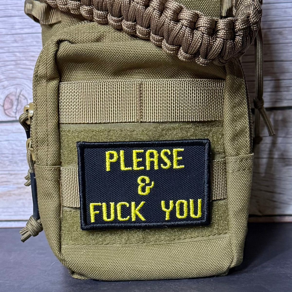 A close-up of a tactical pouch in olive drab green nylon with a black rectangular patch featuring yellow embroidered text that reads 'PLEASE & FUCK YOU'. A tan paracord strap is threaded through the top zipper, and the pouch is photographed against a rustic wood plank background.