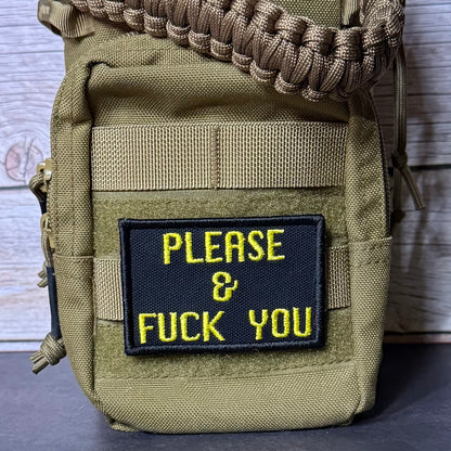 A close-up of a tactical pouch in olive drab green nylon with a black rectangular patch featuring yellow embroidered text that reads 'PLEASE & FUCK YOU'. A tan paracord strap is threaded through the top zipper, and the pouch is photographed against a rustic wood plank background.