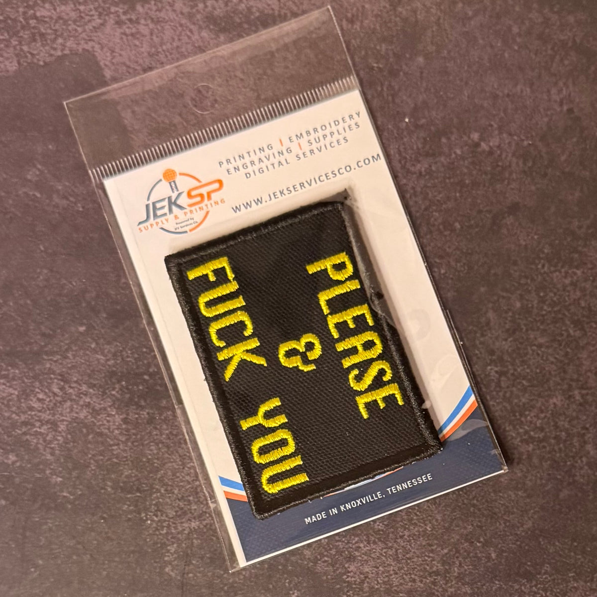 A black rectangular embroidered patch with yellow text reading 'PLEASE & FUCK YOU' inside a clear plastic sleeve, mounted on a branded white backing card. The backing card shows the JEKSP Professional logo and text including 'Printing | Embroidery | Engraving | Supplies | Digital Services' and 'Made in Nashville, Tennessee.