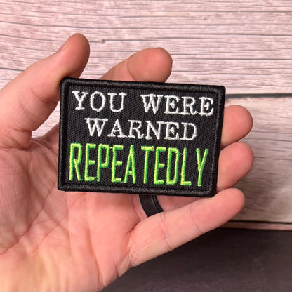A hand holding a rectangular embroidered patch with the text 'YOU WERE WARNED REPEATEDLY' in white and bright green on a black background, set against a light wood plank surface.