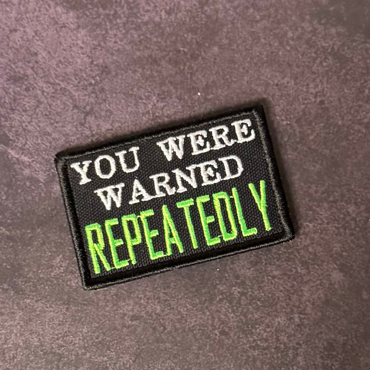 A rectangular embroidered patch with a black background and the text 'YOU WERE WARNED REPEATEDLY' in white and bright green lettering, placed on a textured dark gray surface.