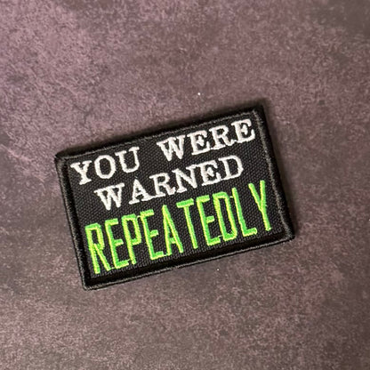 A rectangular embroidered patch with a black background and the text 'YOU WERE WARNED REPEATEDLY' in white and bright green lettering, placed on a textured dark gray surface.