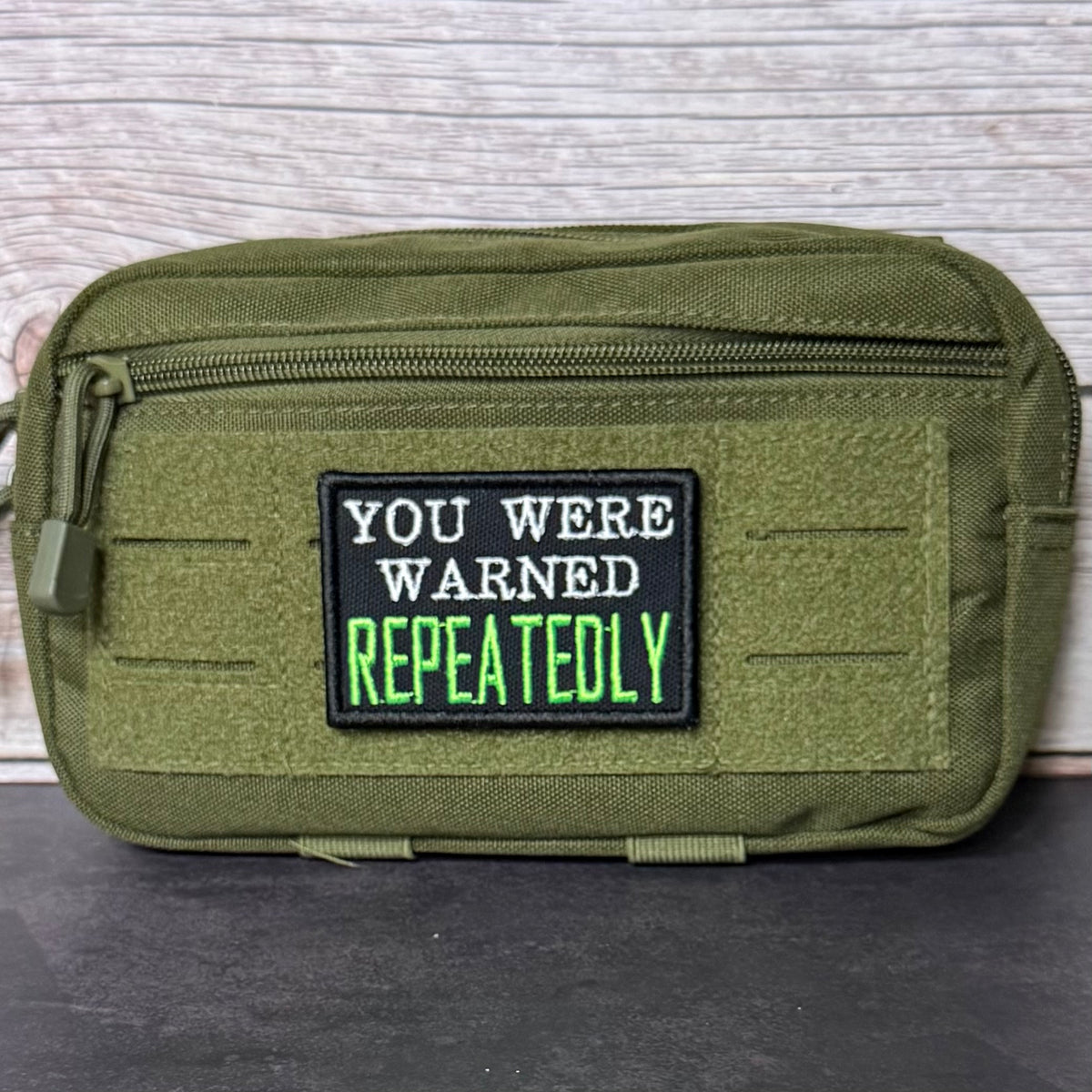 An olive green tactical pouch with a black rectangular patch that reads 'YOU WERE WARNED REPEATEDLY' in white and neon green text, set against a light wood panel background.