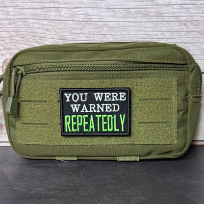An olive green tactical pouch with a black rectangular patch that reads 'YOU WERE WARNED REPEATEDLY' in white and neon green text, set against a light wood panel background.