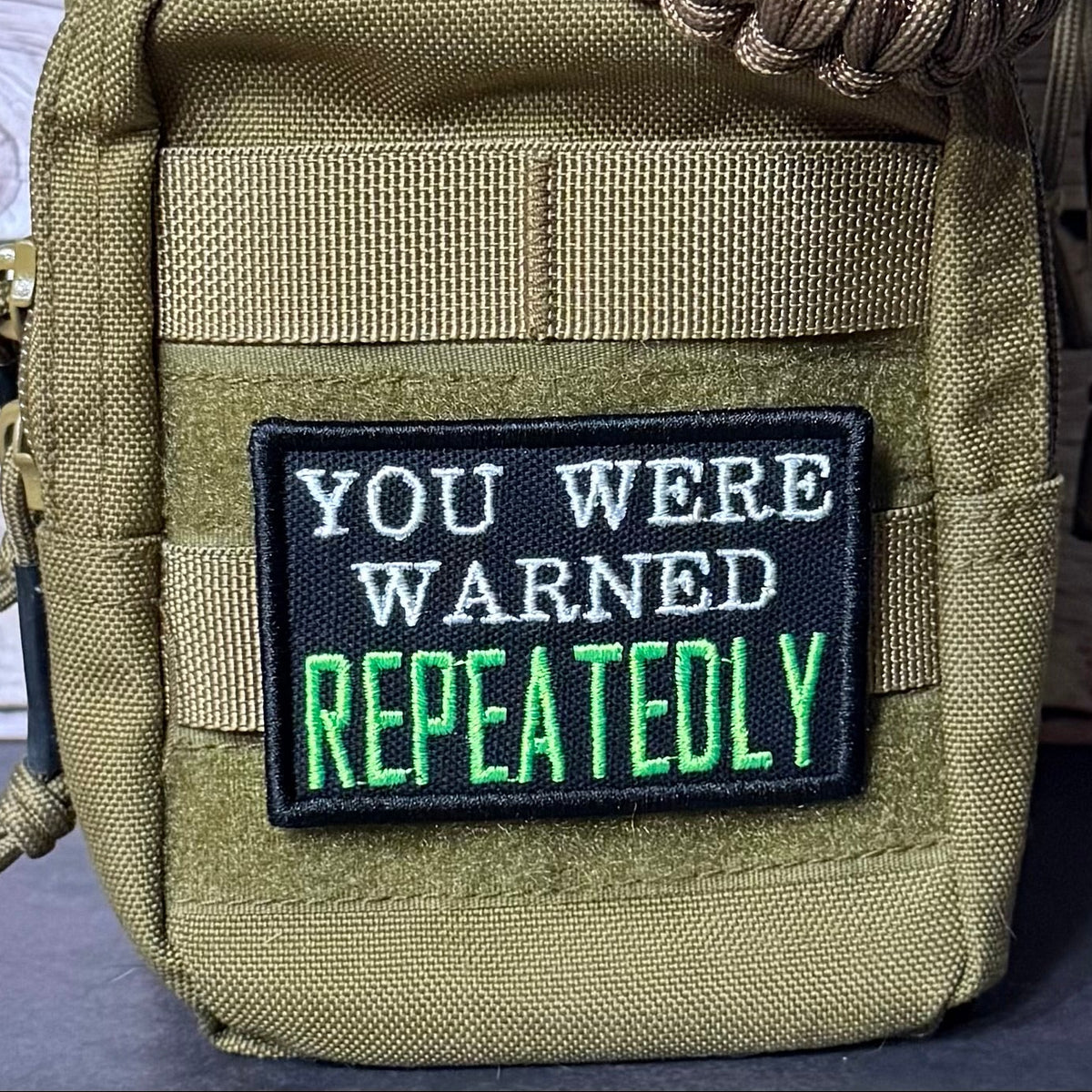A tactical pouch in olive drab nylon with a black embroidered patch that reads 'YOU WERE WARNED REPEATEDLY' in white and green text, shown close-up with a paracord strap.