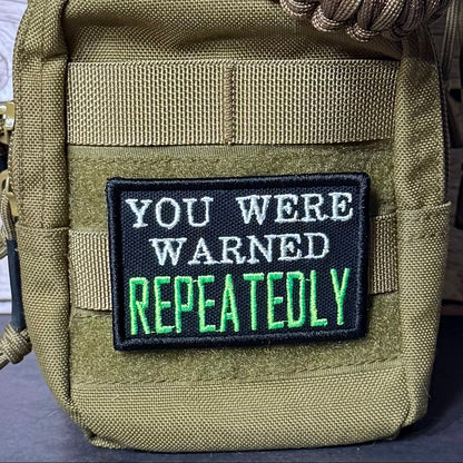 A tactical pouch in olive drab nylon with a black embroidered patch that reads 'YOU WERE WARNED REPEATEDLY' in white and green text, shown close-up with a paracord strap.