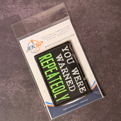 An embroidered patch in clear plastic packaging, featuring a black background with bright green and white text that reads 'REPEATEDLY YOU WERE WARNED'. The packaging card shows the JEKSP brand logo and website.