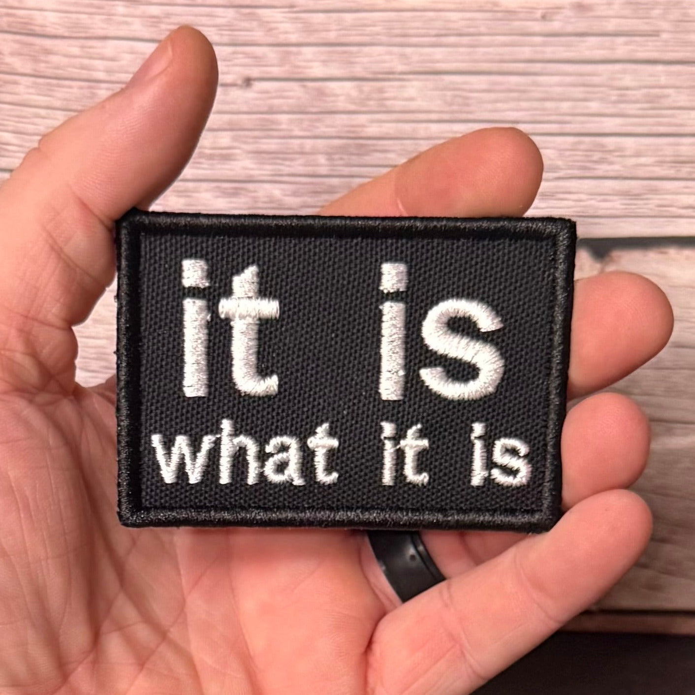 A hand holding a rectangular black embroidered patch with the white text 'it is what it is' against a light wood background.