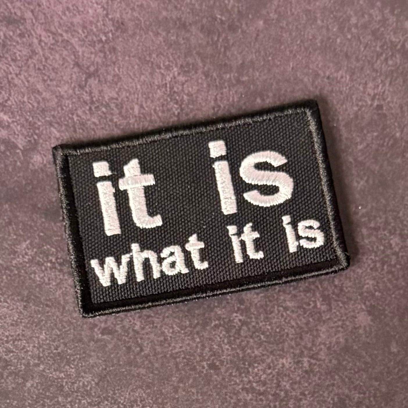 A rectangular black embroidered patch with the phrase 'it is what it is' in white text, placed on a textured mauve-brown fabric background.