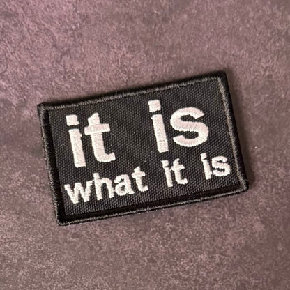 A rectangular black embroidered patch with the phrase 'it is what it is' in white text, placed on a textured mauve-brown fabric background.