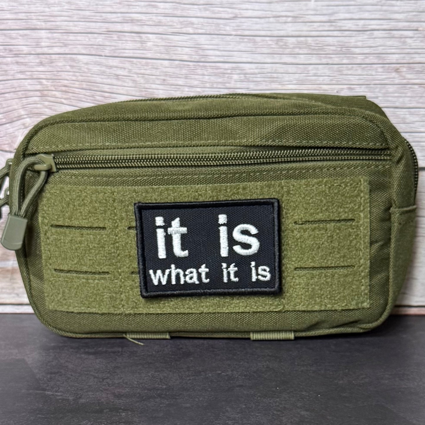 An olive green tactical pouch with a black rectangular patch that reads 'it is what it is' in white text, shown against a light wood background.