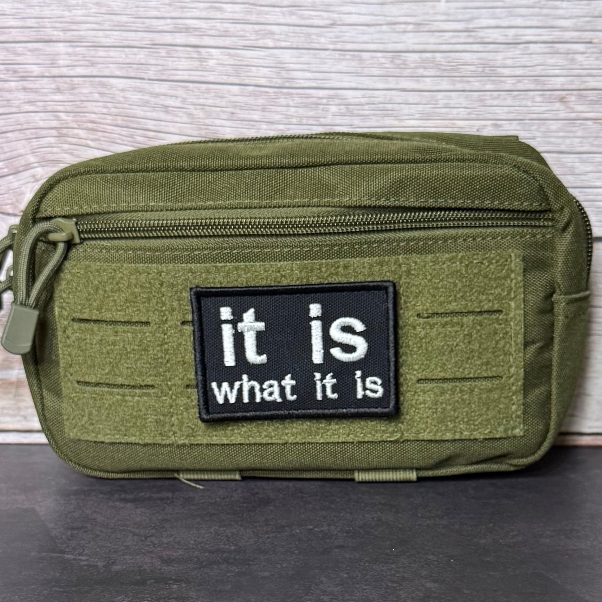An olive green tactical pouch with a black rectangular patch that reads 'it is what it is' in white text, shown against a light wood background.