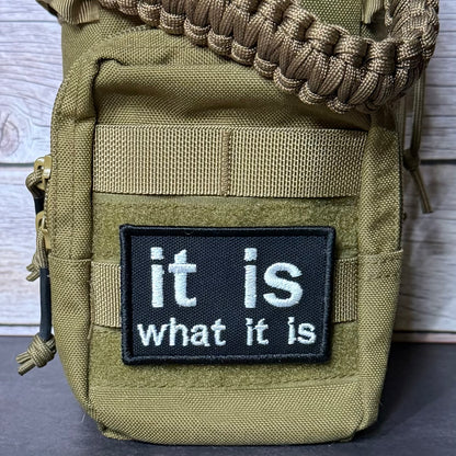 Close-up of a tactical pouch in olive drab green with a black rectangular patch that reads 'it is what it is' in white text, plus a tan paracord strap wrapped around the top.