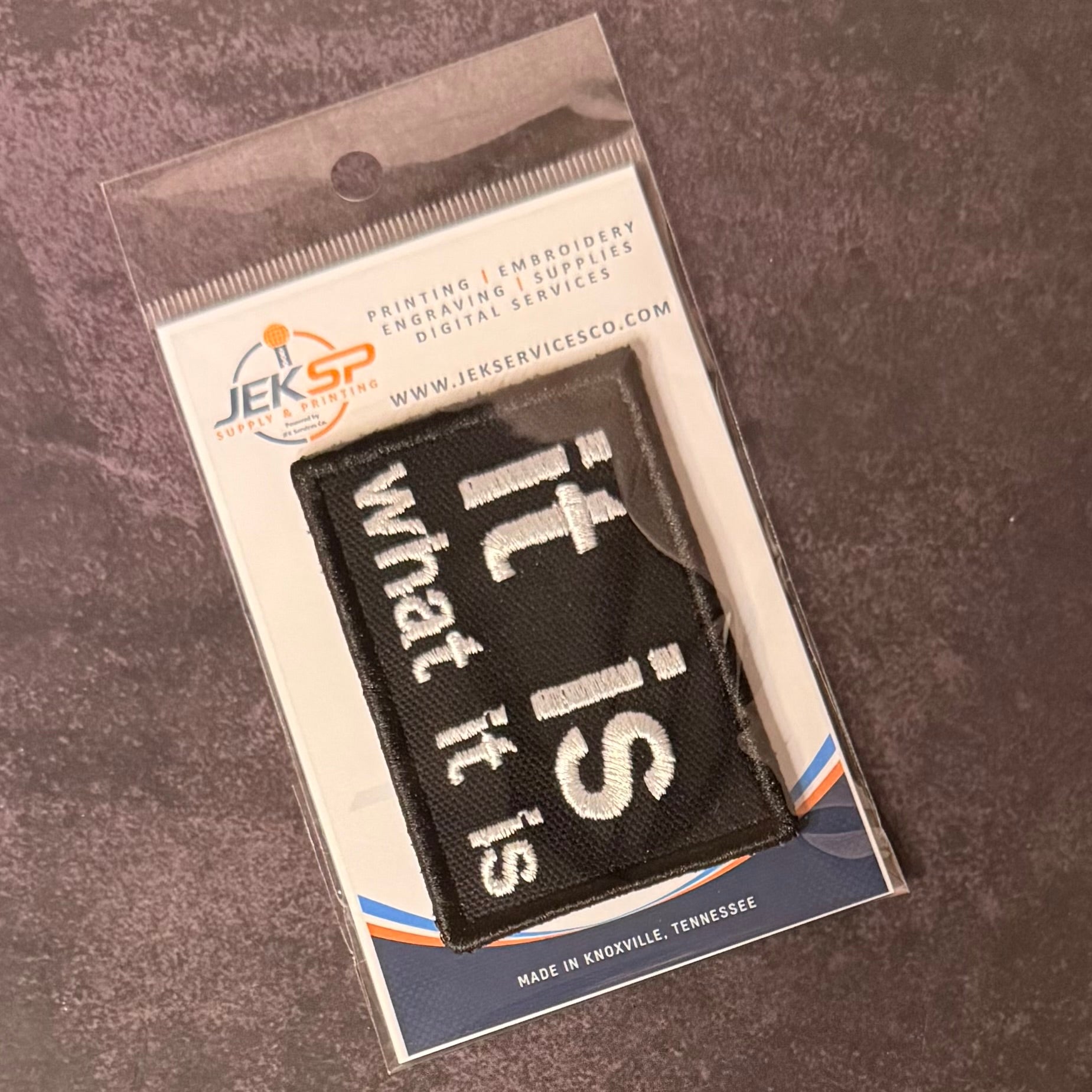 A black rectangular embroidered patch with white text reading 'it is what it is' in a diagonal layout, sealed inside a clear plastic retail package with a printed backing card. The package shows the JEKSP brand and is photographed on a textured dark gray surface.