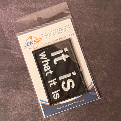 A black rectangular embroidered patch with white text reading 'it is what it is' in a diagonal layout, sealed inside a clear plastic retail package with a printed backing card. The package shows the JEKSP brand and is photographed on a textured dark gray surface.