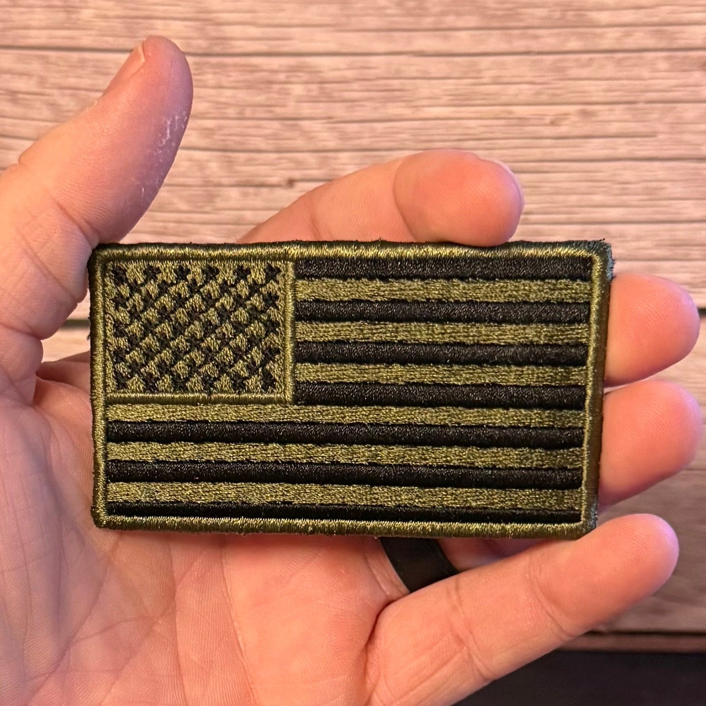 An olive drab and black embroidered American flag patch held in a person's hand against a light wood background.