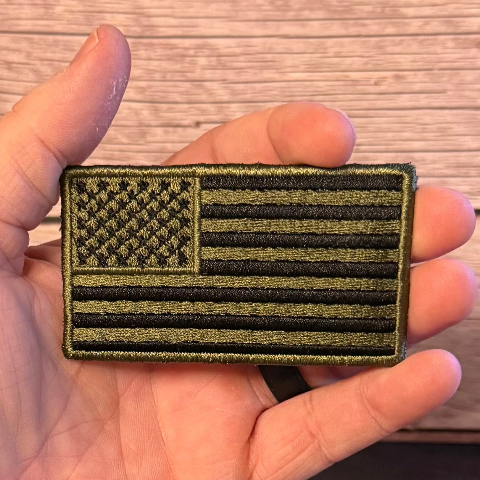 An olive drab and black embroidered American flag patch held in a person's hand against a light wood background.