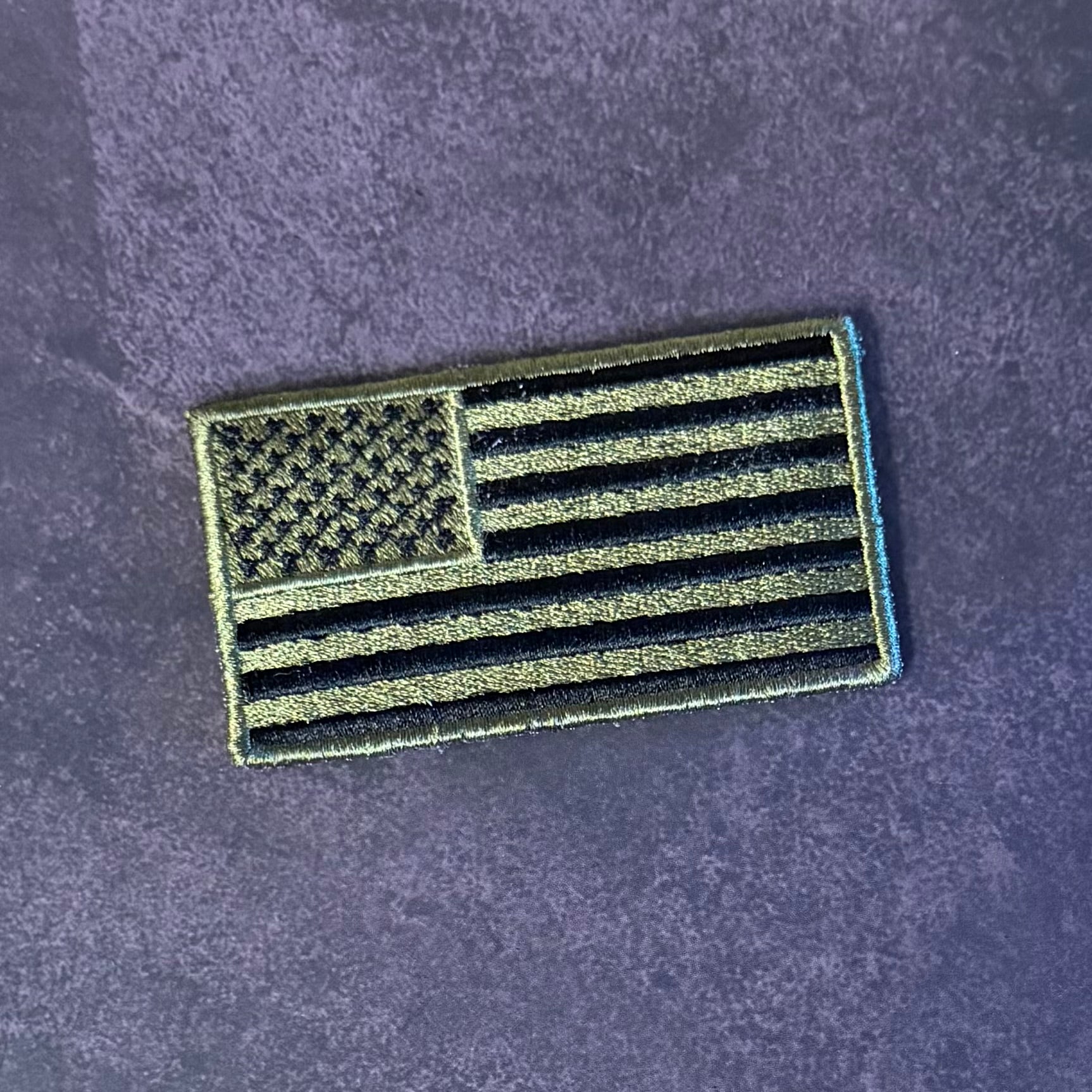 An embroidered United States flag patch in olive drab and black, displayed on a textured dark purple background.
