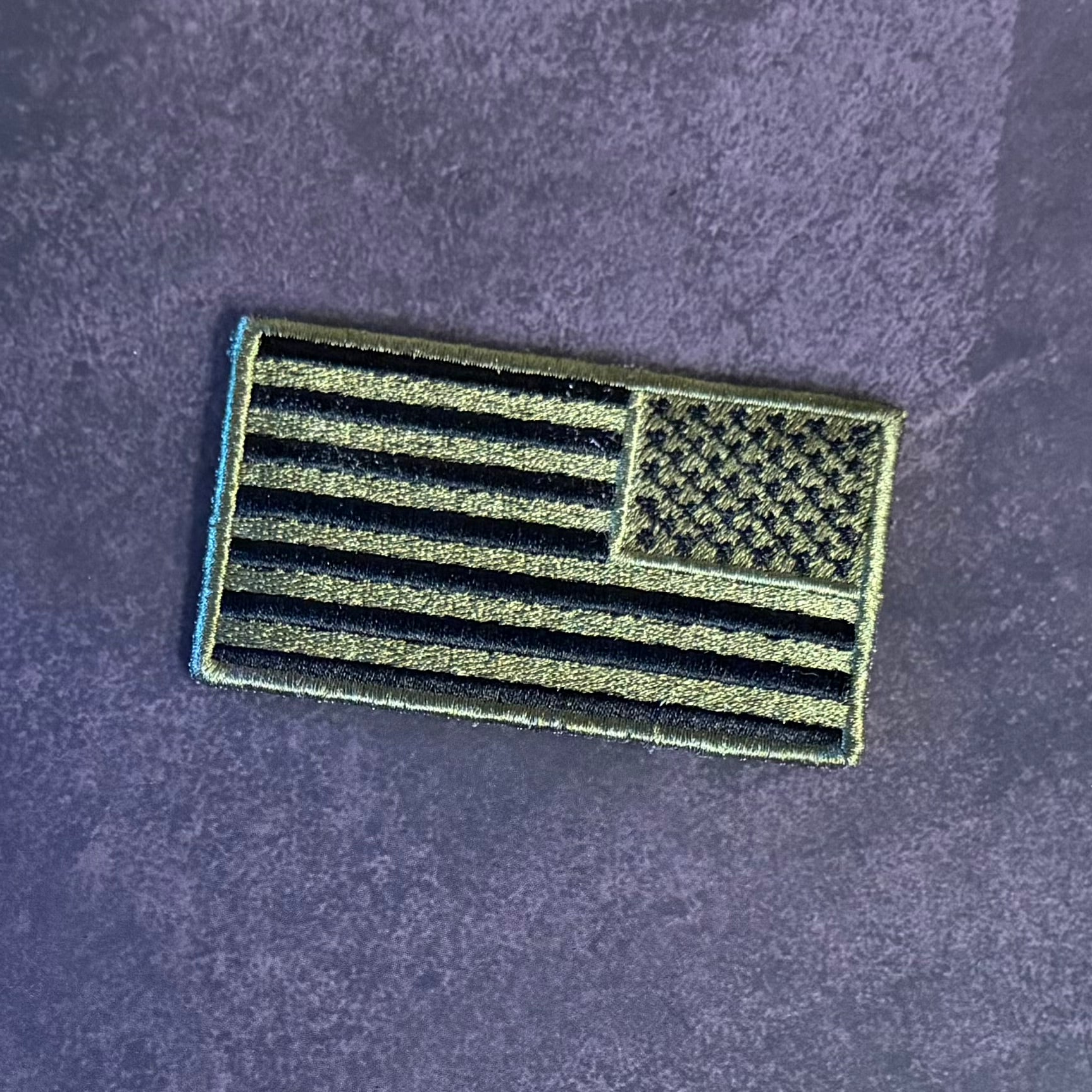 An embroidered American flag patch with black and olive drab stripes and canton, shown on a textured dark purple background.